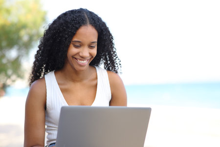 Happy black woman checking laptop sitting on the beachの写真素材