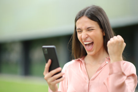 Excited elegant woman celebrating success holding phone outdoorsの写真素材