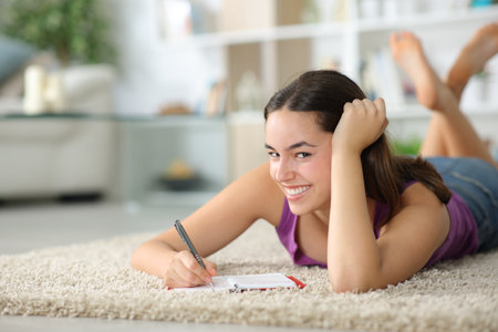 Happy woman lying on a carpet at home writing in agenda looking at youの写真素材
