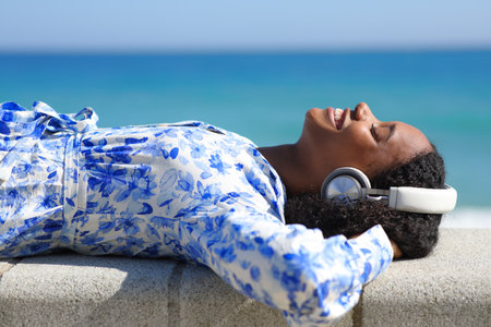 Happy black woman wearing headphone lying listening music on the beachの写真素材