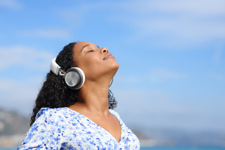 Black woman with headphone breathing fresh air listening guide on the beach a sunny dayの写真素材