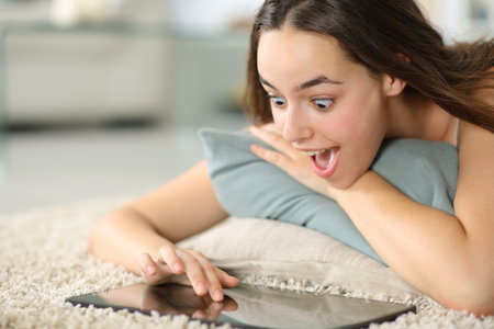 Amazed woman checking tablet lying on the floor at homeの写真素材