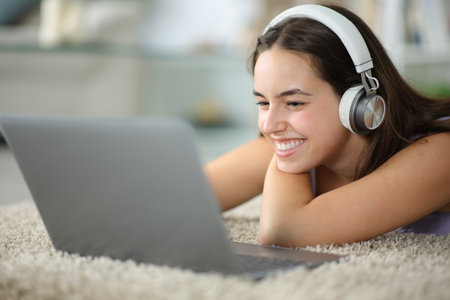 Happy woman with headphone checks laptop lying on the floor at homeの写真素材