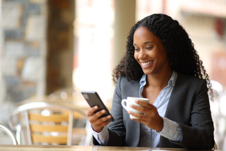 Happy black executive drinking and using phone in a restaurant terraceの写真素材