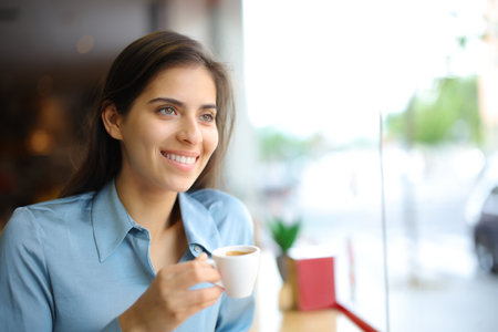 Happy elegant woman smiling drinking coffee in a bar interior looking through a windowの写真素材