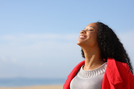 Happy black woman wearing red jacket breathing fresh air on the beach in winter a sunny dayの写真素材