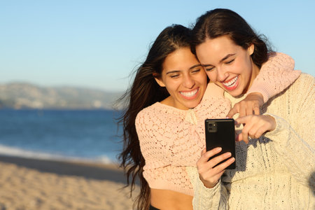 Two joyful friends checking smart phone on the beach at sunset