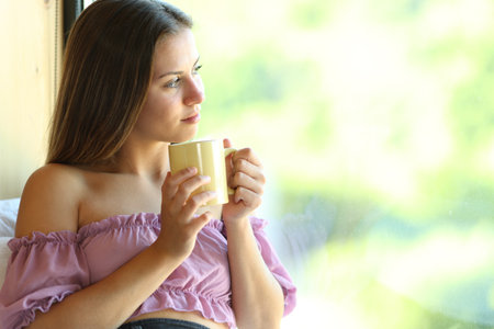 Teen looking through a window drinking tea at homeの写真素材