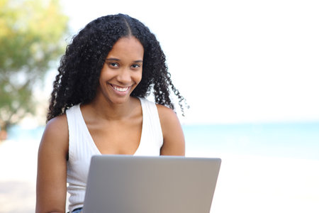 Happy black woman looks at you with a laptop sitting on the beachの写真素材