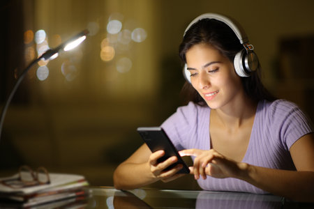 Happy woman in the night using phone wearing headphone at homeの写真素材