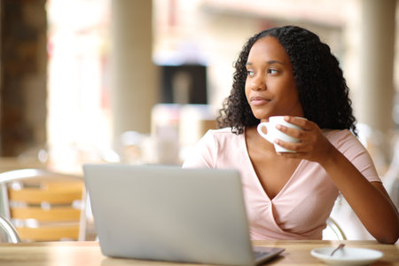 Black serious woman in a bar with a laptop drinking and looking awayの写真素材