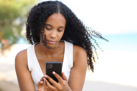 Front view portrait of a black serious woman using cell phone on the beachの写真素材