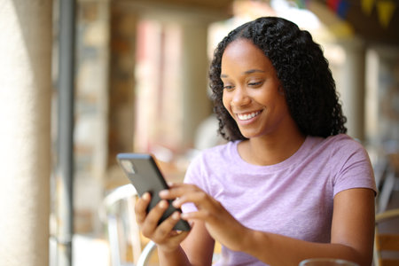 Happy black woman is using smart phone in a restaurant terraceの写真素材