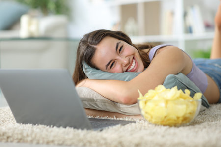 Happy woman with laptop and snack looking at you lying on a carpet at homeの写真素材