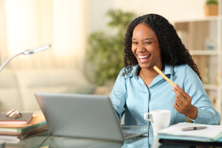 Happy black businesswoman eating snack using laptop at homeの写真素材