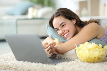 Happy woman watching movie eating potato chips lying on the floor at homeの写真素材