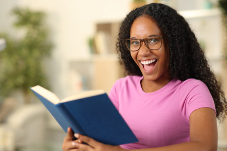 Happy black reader with eyeglasses looks at you holding a paper book at homeの写真素材