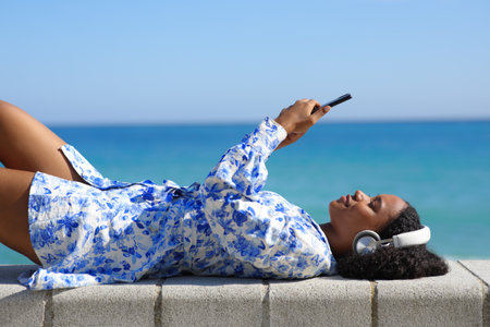 Profile of a happy black woman wearing headphone watching media on phone lying on the beachの写真素材