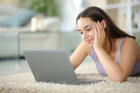 Satisfied woman lying on the floor watching videos on laptop at homeの写真素材