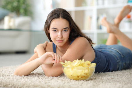 Beautiful woman looks at you eating potato chips lying on a carpet at homeの写真素材