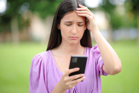 Front view portrait of a worried woman checking bad news on smart phone in a parkの写真素材