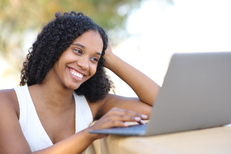Happy black woman using laptop watching online content sitting outdoorsの写真素材