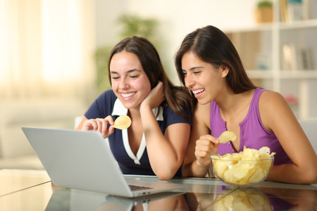 Two happy friends watching movie on laptop eating snack sitting at homeの写真素材