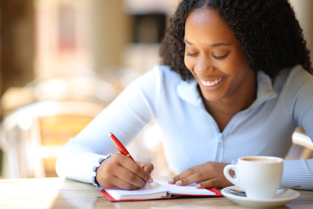 Happy black woman writing in agenda sitting in a coffee shop terraceの写真素材