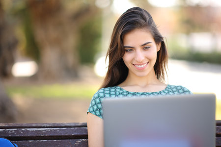 Front view portrait of a happy student using laptop sitting in a parkの写真素材