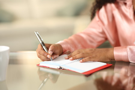 Close up of a black woman hands writing in paper agenda at homeの写真素材
