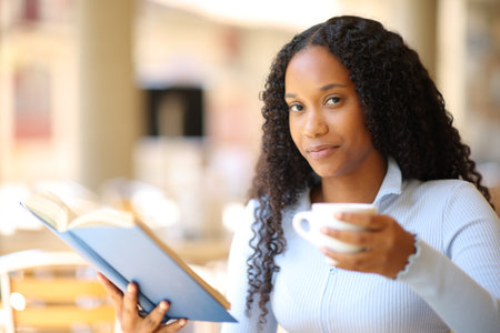 Happy black woman holding paper book and coffee cup looking at you in a restaurant terraceの写真素材