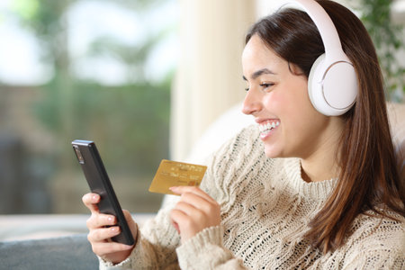 Happy woman with headphone buying media online with credit card and smart phone at homeの写真素材