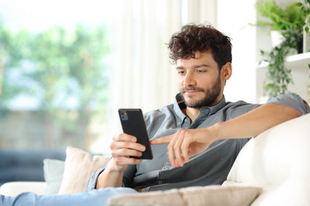 Serious man using smart phone sitting on a couch at homeの写真素材
