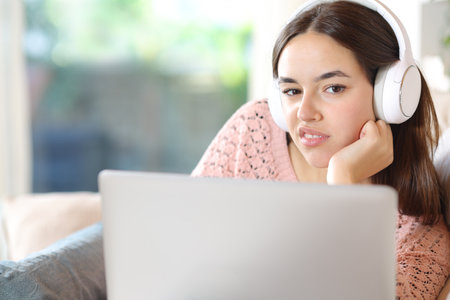 Disgusted woman with headphone and laptop looks at you sitting at homeの写真素材