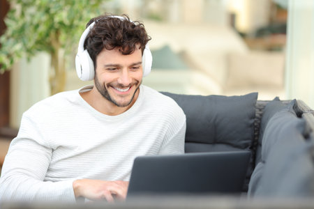 Happy man wearing headphone watching media on laptop sitting on a couch in a house terraceの写真素材