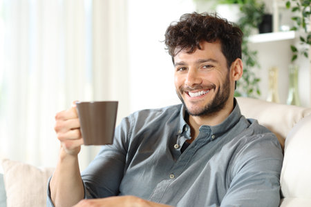 Happy man drinking coffee sitting on a couch at home looking at cameraの写真素材