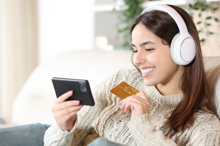 Happy woman wearing headphone buying media products with credit card and phone at homeの写真素材