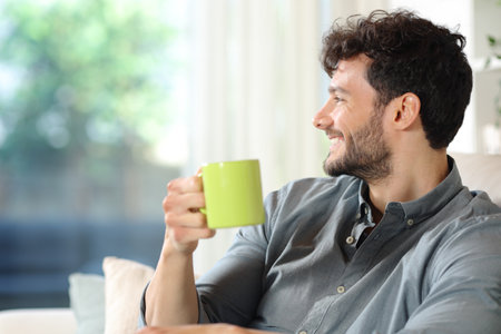 Profile of a happy man drinking coffee looking through a window sitting on a couch at homeの写真素材