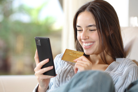 Happy woman buying online with phone and credit card sitting on a couch at homeの写真素材