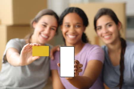 Three happy multi ethnic roommates moving home showing blank phone screen and credit card