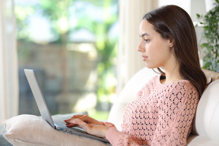 Profile of a serious woman using laptop sitting on a couch at homeの写真素材