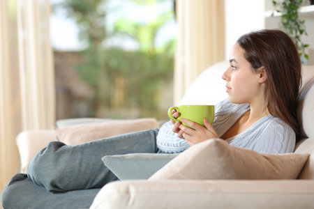 Pensive woman relaxing drinking coffee looking at garden through a window at homeの写真素材