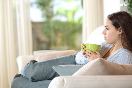 Serious pensive woman drinking coffee lying on a couch at homeの写真素材