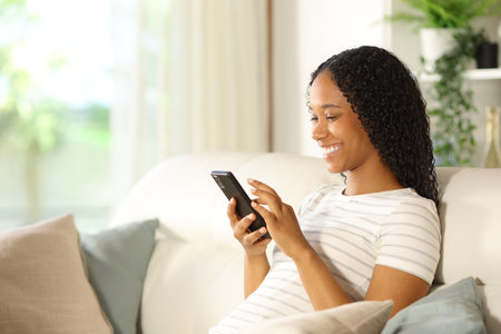Happy black woman using smart phone sitting on a couch at homeの写真素材