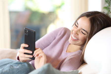 Happy woman sitting on a couch checking smart phone and smilingの写真素材