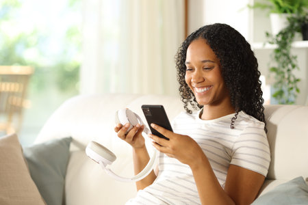 Happy black woman pairing wireless headphone with smart phone sitting on a couch at homeの写真素材