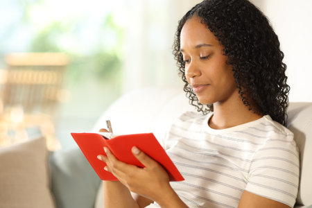 Serious black woman writing in red agenda at homeの写真素材