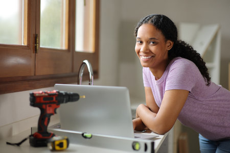 Happy black woman with a laptop renovating house looks at you in the kitchenの写真素材