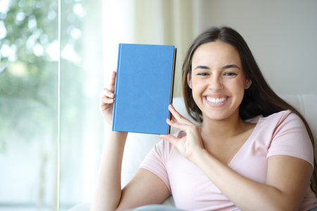Happy woman showing at camera blank paper book cover at homeの写真素材