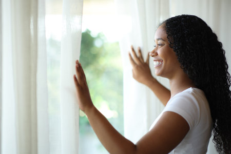 Happy black woman opening curtains and looking through a window at homeの写真素材